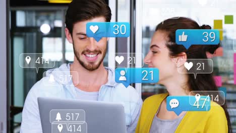 Animation-of-social-media-icons-over-diverse-man-and-woman-smiling-looking-at-the-laptop-at-office