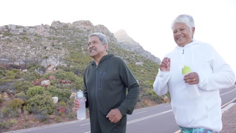 Happy-senior-biracial-couple-running-on-road-with-bottles-of-water-in-mountains