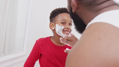 Happy-african-american-father-and-son-applying-shaving-cream-on-face-in-bathroom,-in-slow-motion