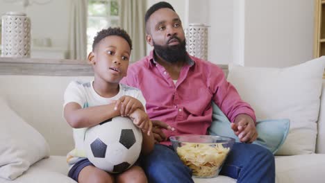 Glücklicher-Afroamerikanischer-Vater-Und-Sohn,-Die-Sich-Ein-Fußballspiel-Ansehen-Und-Chips-Essen,-In-Zeitlupe