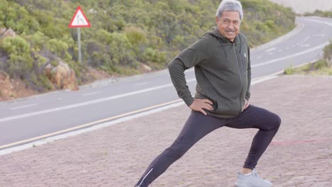 Happy-senior-biracial-man-stretching-at-road-in-mountains