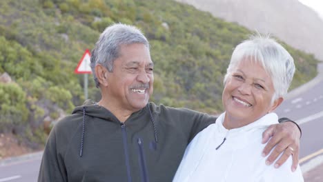 Portrait-of-happy-senior-biracial-couple-embracing-at-road-in-mountains