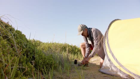 Happy-senior-biracial-man-preparing-tent-in-mountains-on-sunny-day,-in-slow-motion