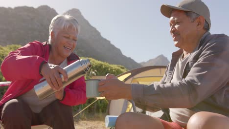 Happy-senior-biracial-couple-sitting-at-tent-in-mountains-and-drinking-coffee,-in-slow-motion
