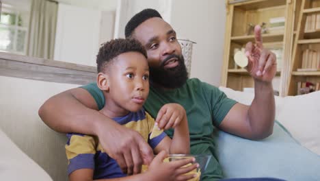 Glücklicher-Afroamerikanischer-Vater-Und-Sohn-Sitzen-Auf-Dem-Sofa,-Schauen-Fern-Und-Essen-Chips,-In-Zeitlupe