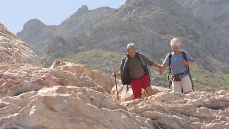 Happy-senior-biracial-couple-in-mountains-hiking-with-trekking-sticks,-in-slow-motion