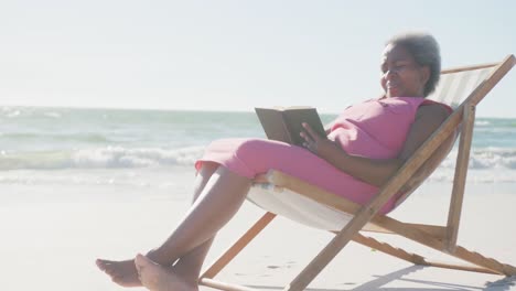 Glückliche-ältere-Afroamerikanische-Frau-Sitzt-Auf-Einem-Liegestuhl-Und-Liest-Ein-Buch-Am-Strand,-In-Zeitlupe