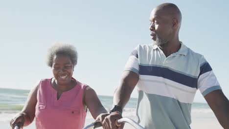 Glückliches-älteres-Afroamerikanisches-Paar,-Das-Mit-Fahrrädern-Am-Strand-Spaziert,-In-Zeitlupe
