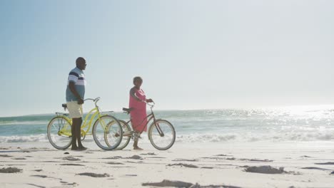 Happy-senior-african-american-couple-walking-with-bikes-at-beach,-copy-space,-in-slow-motion