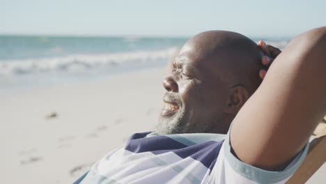 Happy-senior-african-american-man-sitting-on-deck-chair-at-beach,-in-slow-motion