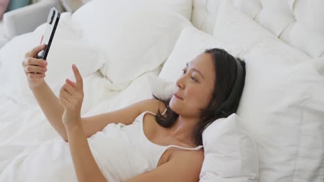 Feliz-Mujer-Asiática-Acostada-En-La-Cama,-Usando-Auriculares-Y-Usando-Un-Teléfono-Inteligente,-En-Cámara-Lenta