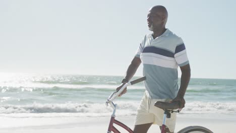 Happy-senior-african-american-man-walking-with-bike-at-beach,-in-slow-motion