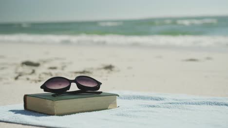Close-up-of-book,-sunglasses-and-towel-on-beach,-in-slow-motion,-with-copy-space