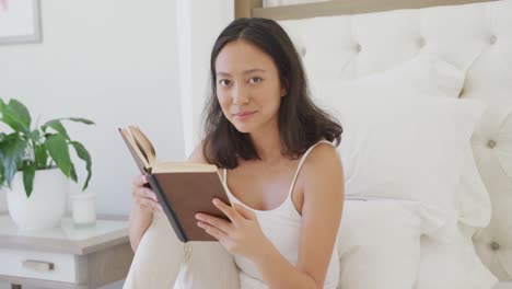 Portrait-of-happy-asian-woman-sitting-on-bed-and-reading-book,-in-slow-motion