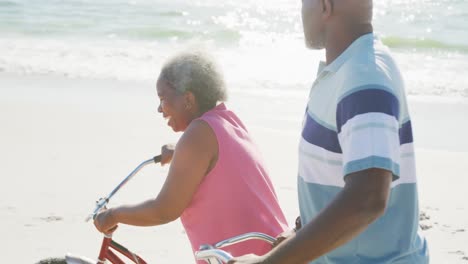 Happy-senior-african-american-couple-walking-with-bikes-at-beach,-in-slow-motion