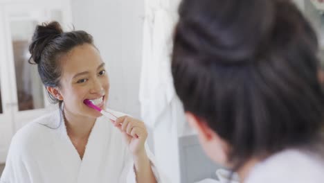 Happy-asian-woman-looking-at-mirror-and-brushing-teeth-in-bathroom,-in-slow-motion