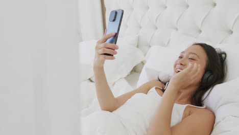 Happy-asian-woman-lying-in-bed,-wearing-headphones-and-using-smartphone,-in-slow-motion