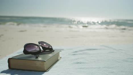 Close-up-of-book,-sunglasses-and-towel-on-beach,-in-slow-motion,-with-copy-space