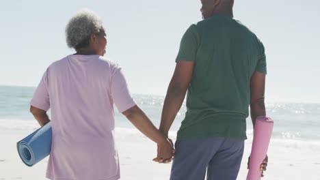 Happy-senior-african-american-couple-walking,-holding-hands-and-yoga-mats-at-beach,-in-slow-motion