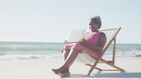 Happy-senior-african-american-woman-sitting-on-deck-chair-and-using-laptop-at-beach,-in-slow-motion