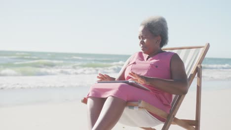 Happy-senior-african-american-woman-sitting-on-deck-chair-at-beach,-in-slow-motion