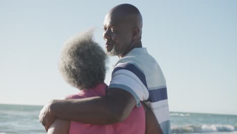 Happy-senior-african-american-couple-walking-and-embracing-at-beach,-in-slow-motion