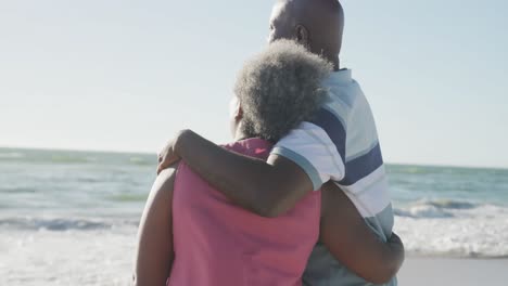 Happy-senior-african-american-couple-walking-and-embracing-at-beach,-in-slow-motion