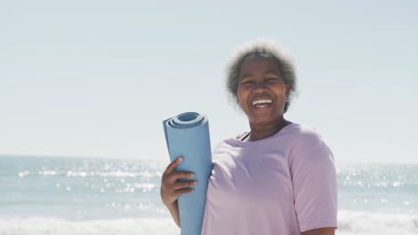 Portrait-of-happy-senior-african-american-woman-holding-yoga-mat-at-beach,-in-slow-motion