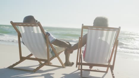 Happy-senior-african-american-couple-sitting-on-deck-chairs,-holding-hands-at-beach,-in-slow-motion
