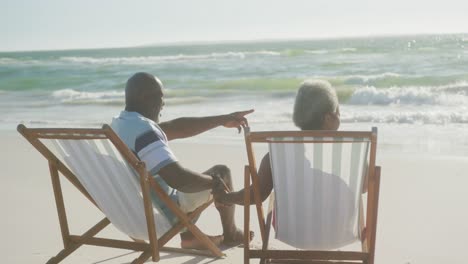 Happy-senior-african-american-couple-sitting-on-deck-chairs,-holding-hands-at-beach,-in-slow-motion