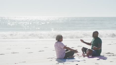 Happy-senior-african-american-couple-doing-yoga-and-meditating-at-beach,-copy-space,-in-slow-motion