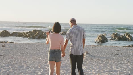 Glückliches-Gemischtrassiges-Paar,-Das-Am-Strand-In-Zeitlupe-Spaziert-Und-Händchen-Hält