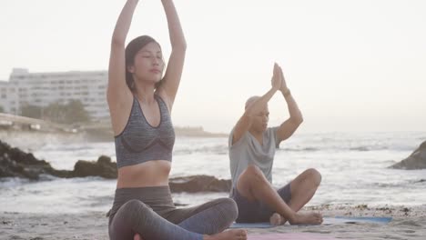 Happy-biracial-couple-doing-yoga-and-meditating-at-beach-at-sundown,-in-slow-motion