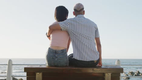 Happy-biracial-couple-sitting-on-bench-and-embracing-on-promenade,-in-slow-motion