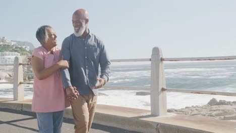 Happy-senior-african-american-couple-walking-along-promenade-by-the-sea,-slow-motion