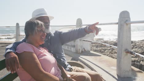 Glückliches-älteres-Afroamerikanisches-Paar-Sitzt-Auf-Einer-Bank-An-Der-Promenade-Am-Meer,-Zeitlupe