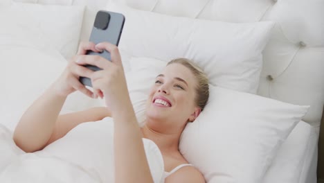 Fröhliche-Kaukasische-Plus-Size-Frau,-Die-In-Zeitlupe-Im-Bett-Lacht-Und-Ihr-Smartphone-Benutzt