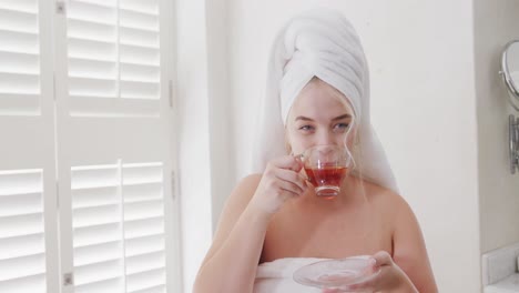 Portrait-of-happy-caucasian-plus-size-woman-with-towel-on-head-drinking-tea-in-slow-motion