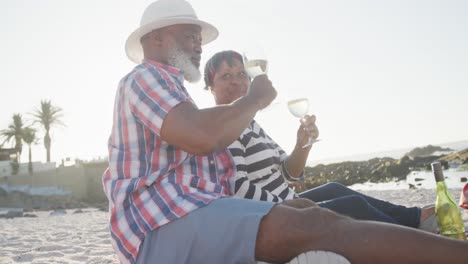 Happy-senior-african-american-couple-having-picnic-at-beach,-drinking-white-wine,-slow-motion