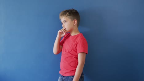 Thoughtful-caucasian-boy-holding-his-chin-on-blue-background,-copy-space,-slow-motion