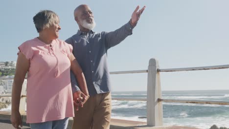 Happy-senior-african-american-couple-walking-along-promenade-by-the-sea,-slow-motion