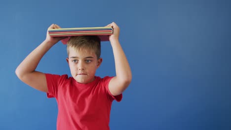 Portrait-of-happy-caucasian-boy-holding-books-on-head-on-blue-background,-copy-space,-slow-motion