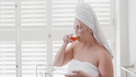 Happy-caucasian-plus-size-woman-with-towel-on-head-drinking-tea-in-slow-motion