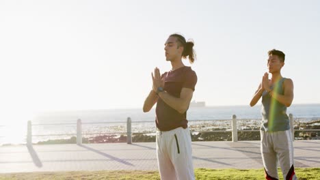 Happy-diverse-gay-male-couple-doing-yoga-and-meditating-at-promenade-by-the-sea,-slow-motion