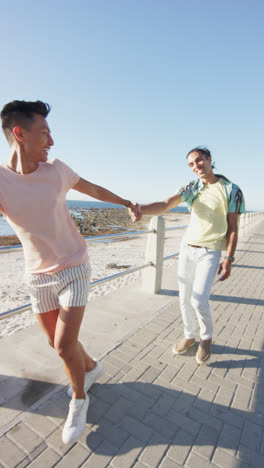 Video-Vertical-De-Una-Feliz-Y-Diversa-Pareja-Masculina-Tomándose-De-La-Mano-En-El-Paseo-Marítimo,-En-Cámara-Lenta