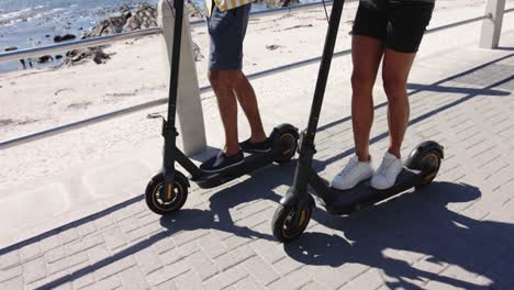 Happy-diverse-gay-male-couple-using-scooters-at-promenade-by-the-sea,-slow-motion
