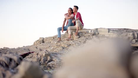 Happy-diverse-gay-male-couple-sitting-on-rocks-and-embracing-at-beach,-slow-motion
