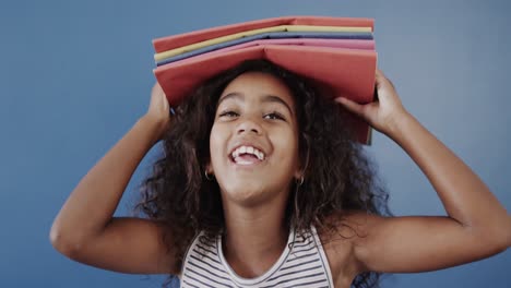 Portrait-of-happy-african-american-girl-holding-books-on-head-on-blue-background,-slow-motion
