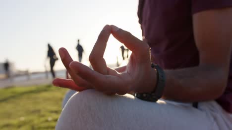 Fröhlicher-Birazialer-Mann,-Der-Yoga-Macht-Und-Meditiert,-Sitzend-Auf-Gras-Am-Meer,-Zeitlupe