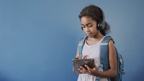 Happy-african-american-girl-using-headphones-and-tablet-on-blue-background,-copy-space,-slow-motion